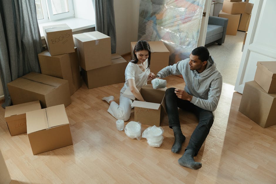 A person sitting on a light wooden floor, holding an open photo album with pictures inside, while another individual kneels beside them, assisting with a cardboard moving box. The box is open and appe